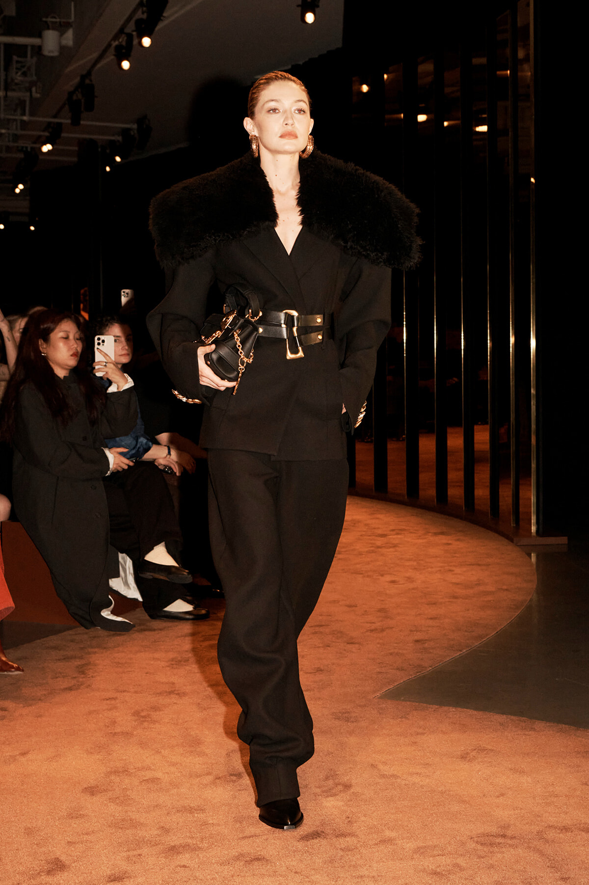 Model on a runway wearing a black tailored suit with an oversized black fur collar, a wide belt, and a small black handbag with a gold chain.