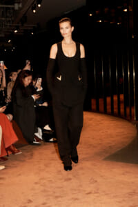 Model walking a beige runway in a black cold-shoulder jumpsuit with pockets, audience taking photos in the background.