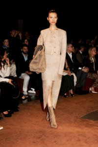 Model walks the runway in a beige suede suit with gold buttons, carrying a large matching bag and wearing knee‑high boots; audience seated along the catwalk behind.