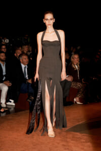 Model on a runway wearing a dark halter-neck gown with long fringe panels flowing from the skirt.