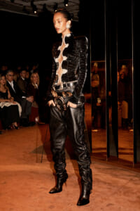 Female model on a runway wearing a black textured leather jacket with white ornate trim, belt, and knee-high boots; audience visible in background.] ,