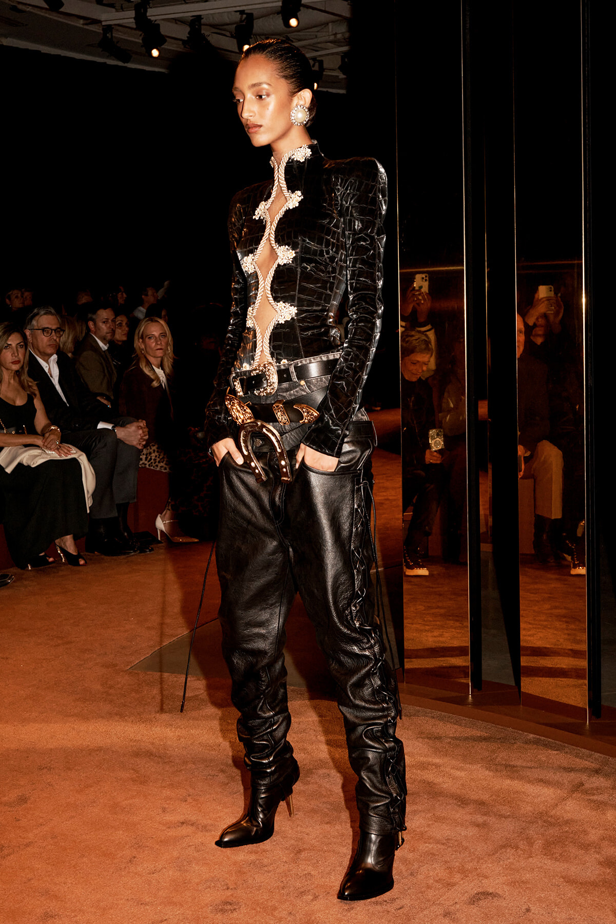 Female model on a runway wearing a black textured leather jacket with white ornate trim, belt, and knee-high boots; audience visible in background.] ,