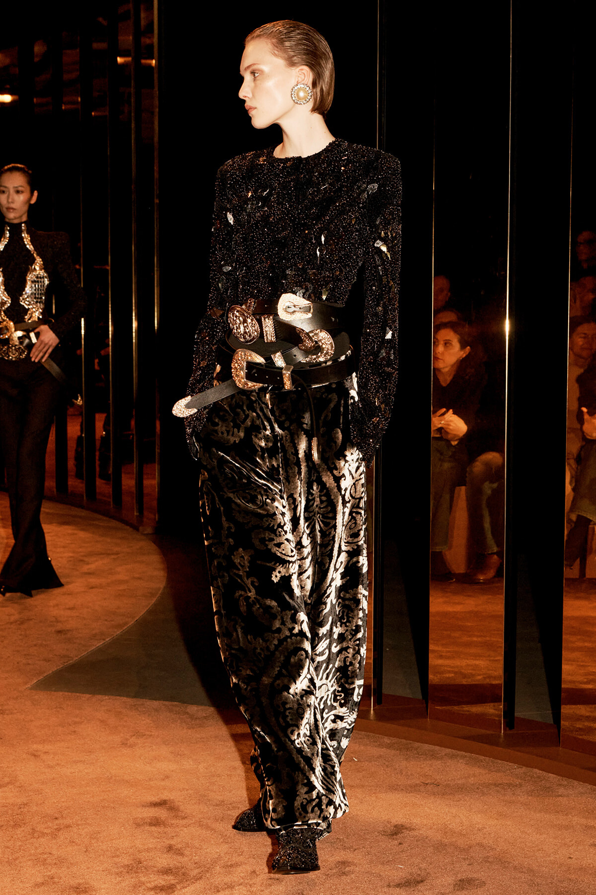 Runway model wearing a black sequined top, ornate stacked belts, and silver brocade pants on a fashion show runway.