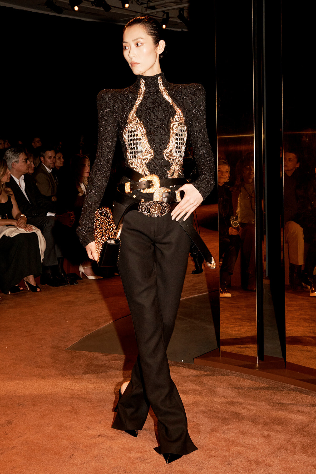 Model on a fashion runway wearing a black sequined turtleneck with gold beaded chest design, black wide-leg pants, and a gold belt with a matching clutch.