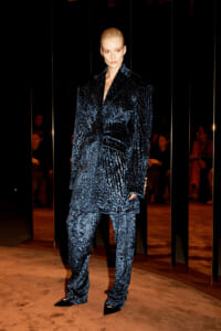 Model wearing a dark blue, heavily textured velvet suit with an oversized blazer and matching wide-leg pants, standing on a runway beside reflective panels.