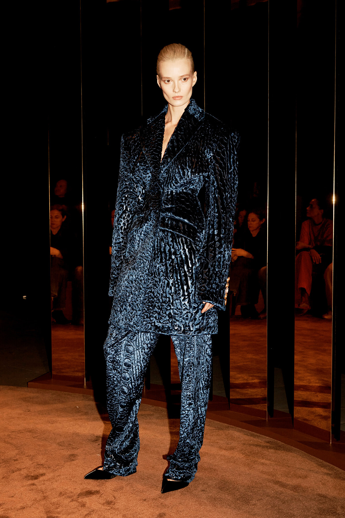 Model wearing a dark blue, heavily textured velvet suit with an oversized blazer and matching wide-leg pants, standing on a runway beside reflective panels.