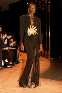 Model walks runway in black fringed gown with large gold sun-shaped emblem, gold hoop earrings, and snakeskin boots; audience watches.