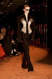 Model walks the runway in a black suit with a white embroidered corset and feather detailing, wearing sunglasses.