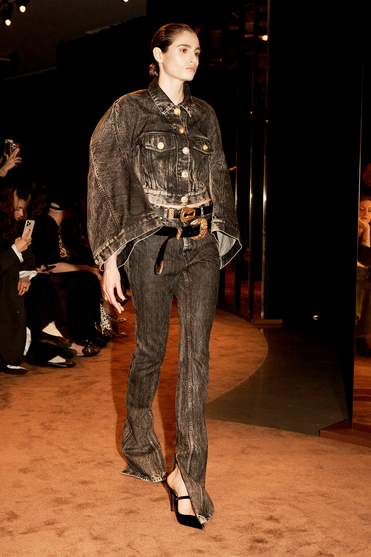 Model on a runway wearing a dark washed denim suit—a cropped jacket with gold buttons and matching jeans, a gold belt, and black heels, with spectators along the catwalk.