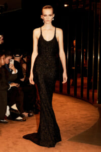 Runway model in a black lace sleeveless gown walking a curved catwalk as audience films with phones nearby.