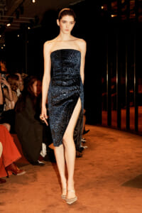 Model walking the runway in a strapless blue textured dress with a high slit, silver heels, and an audience watching.