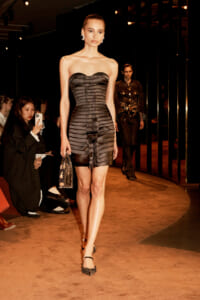 Model walks a runway in a strapless black banded mini dress, holding a small handbag as spectators watch from the sidelines.