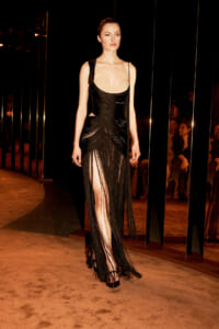 Model walks a runway in a black fringe gown with a long, fringed skirt and high slit on each leg.