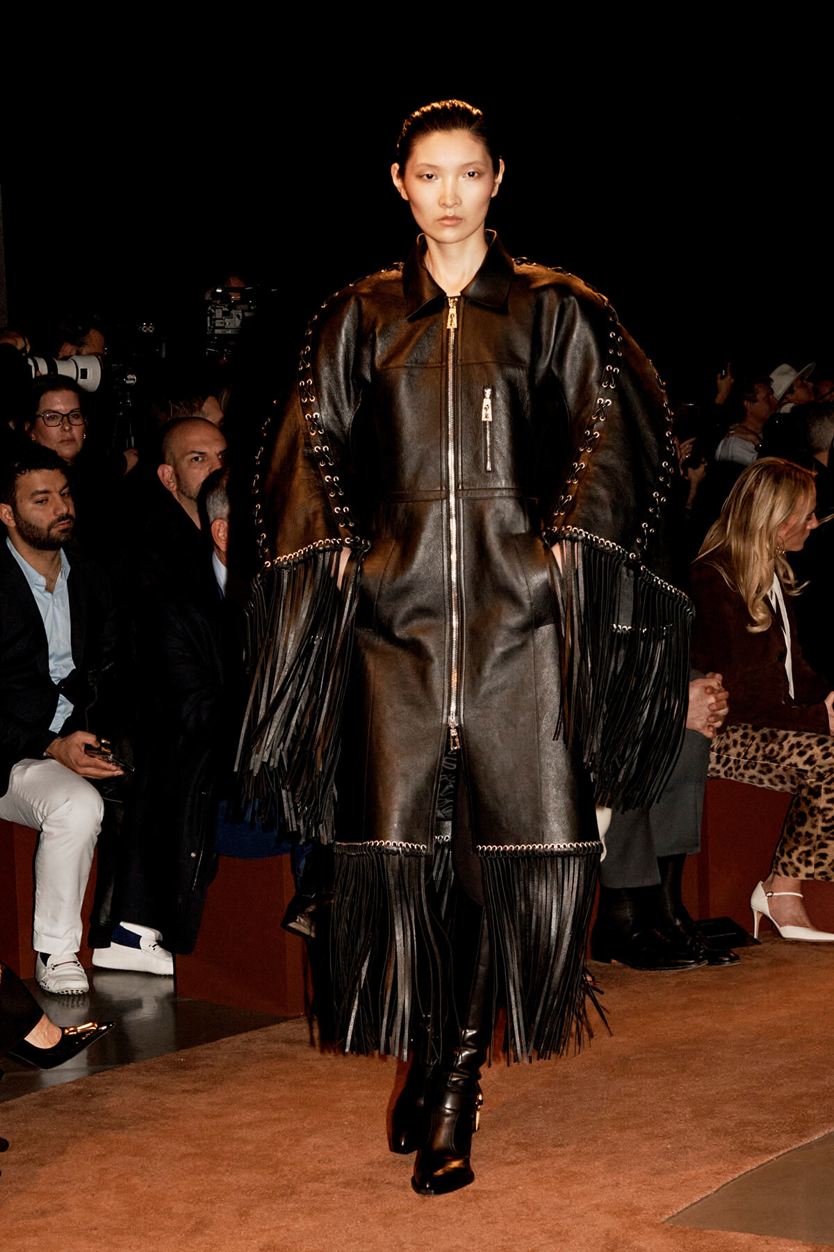 Model walks the runway in a black leather coat with oversized fringe sleeves and laces, audience in background watching.