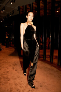 Model walks the runway in a black velvet halter jumpsuit with a gold collar, hands in pockets, under studio lights.