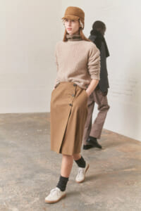 Female model wearing a beige cable-knit turtleneck and tan wrap skirt with buttons, camel cap, and white sneakers.