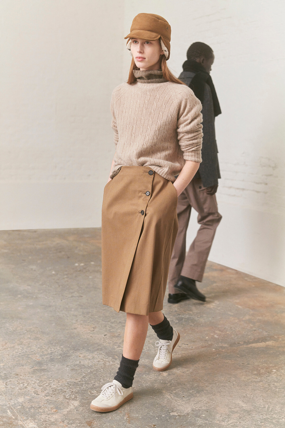 Female model wearing a beige cable-knit turtleneck and tan wrap skirt with buttons, camel cap, and white sneakers.