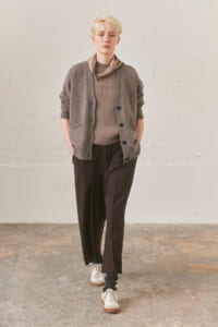 Person with short blond hair wearing a gray cardigan over a brown knit sweater, dark wide-leg pants, a taupe scarf, and white sneakers in a neutral studio setting.