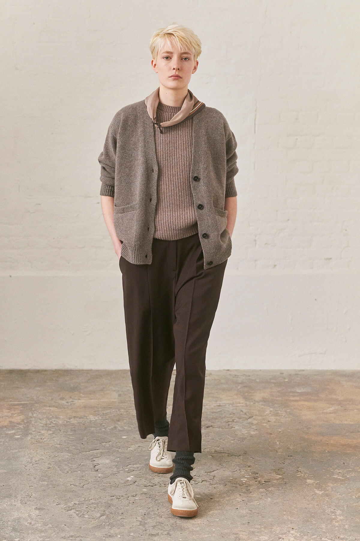 Person with short blond hair wearing a gray cardigan over a brown knit sweater, dark wide-leg pants, a taupe scarf, and white sneakers in a neutral studio setting.