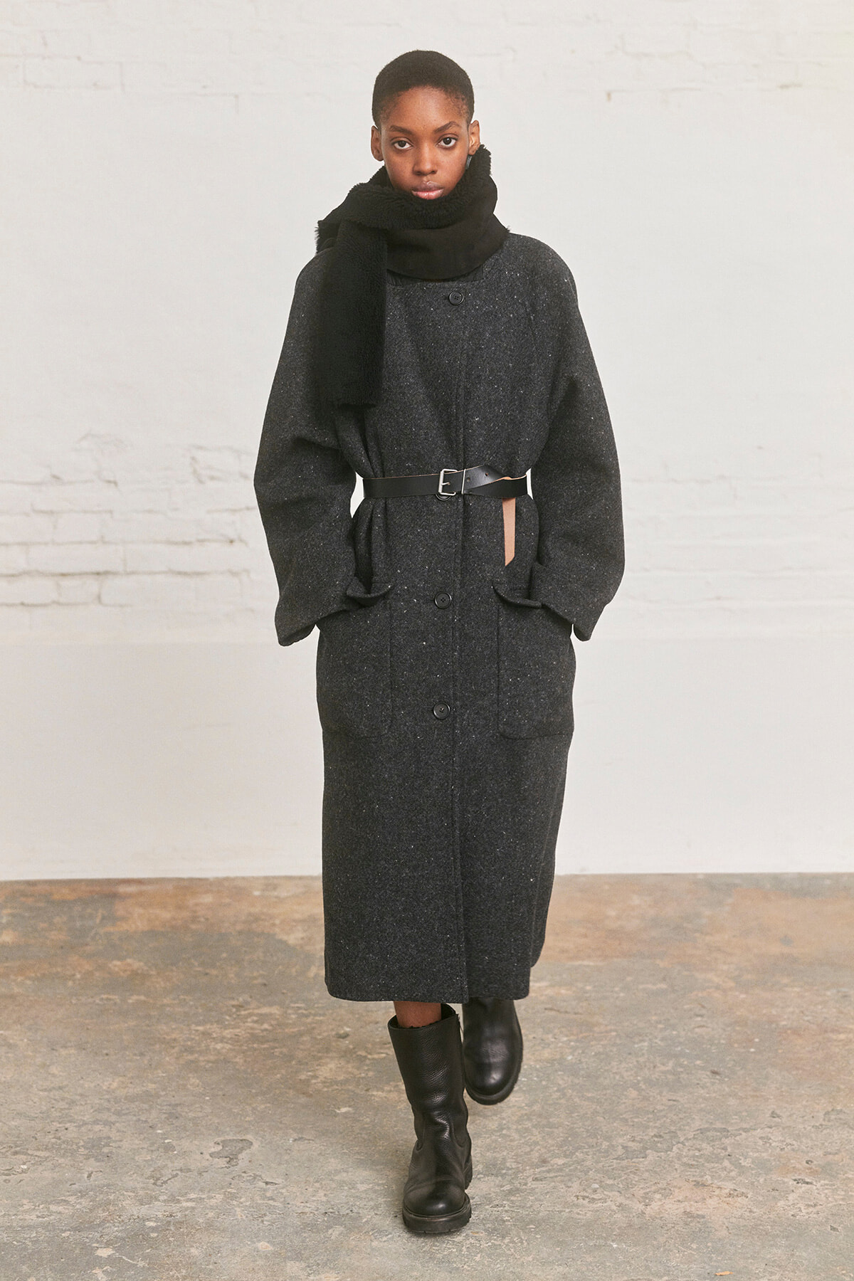 Model wearing a long charcoal-gray coat belted at the waist, with a black scarf and knee-high black boots, standing against a light brick wall.