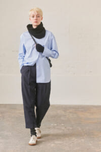 Person with short blond hair wearing an oversized light-blue shirt, navy pants, black scarf and gloves, crossbody bag, and beige sneakers in a studio setting.