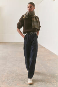 Person standing in a neutral studio wearing a dark brown knit sweater, a plaid scarf, and high-waisted navy trousers with a belt and white sneakers.