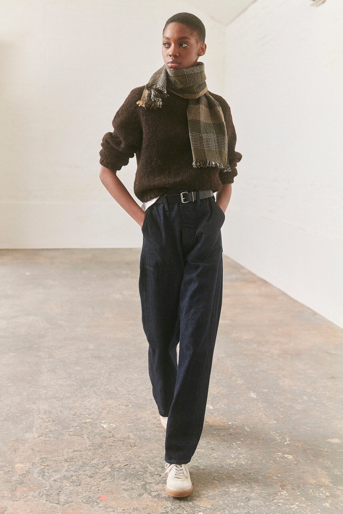 Person standing in a neutral studio wearing a dark brown knit sweater, a plaid scarf, and high-waisted navy trousers with a belt and white sneakers.