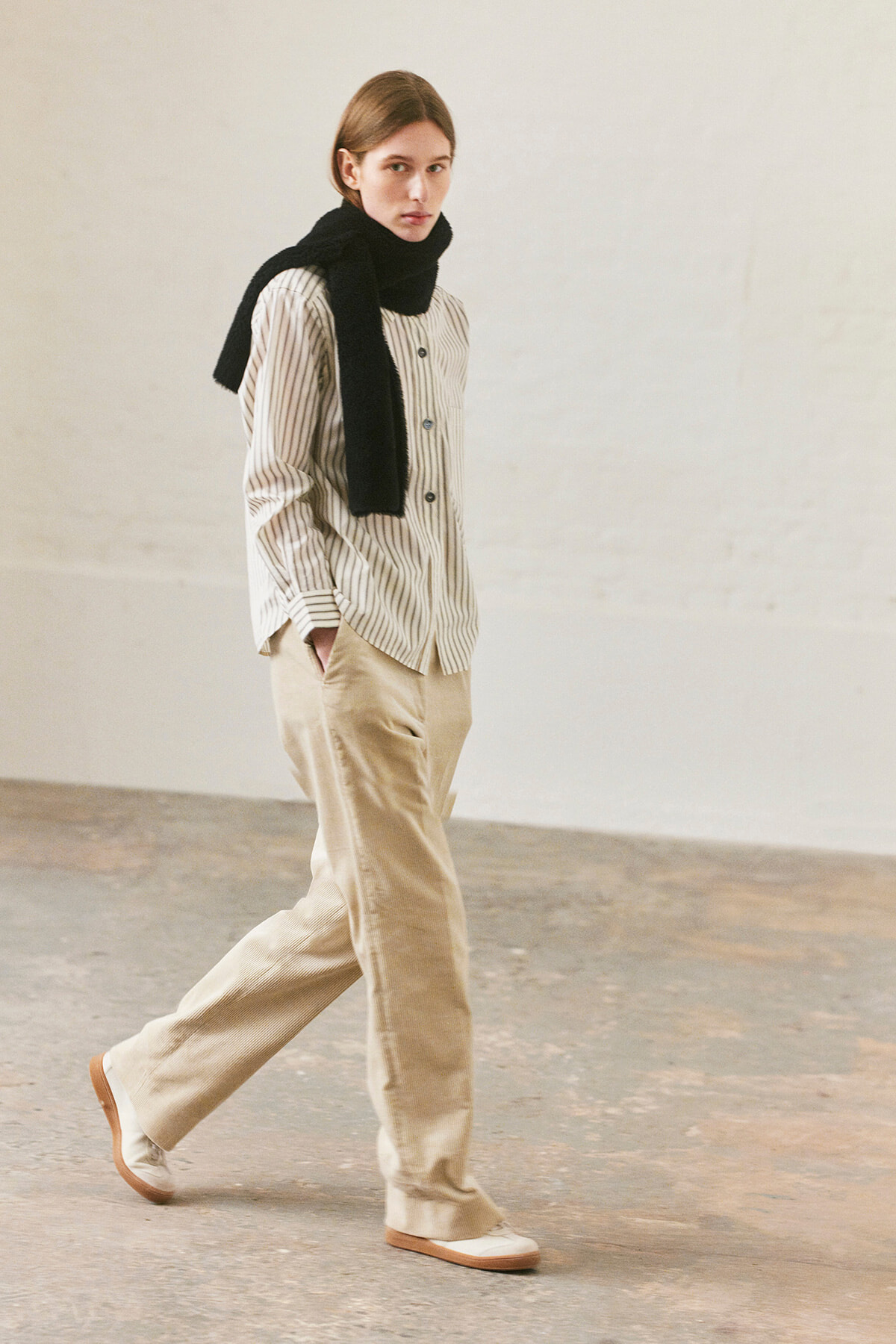 Model walking indoors in a beige-striped shirt, wide-leg beige pants, and a black scarf draped over shoulders.