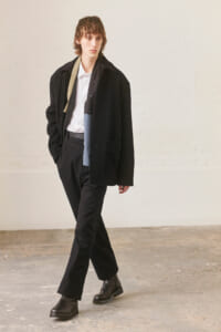 Male model walking in a studio wearing a black overcoat, white shirt, black trousers, black boots, and a beige scarf draped over the shoulder.