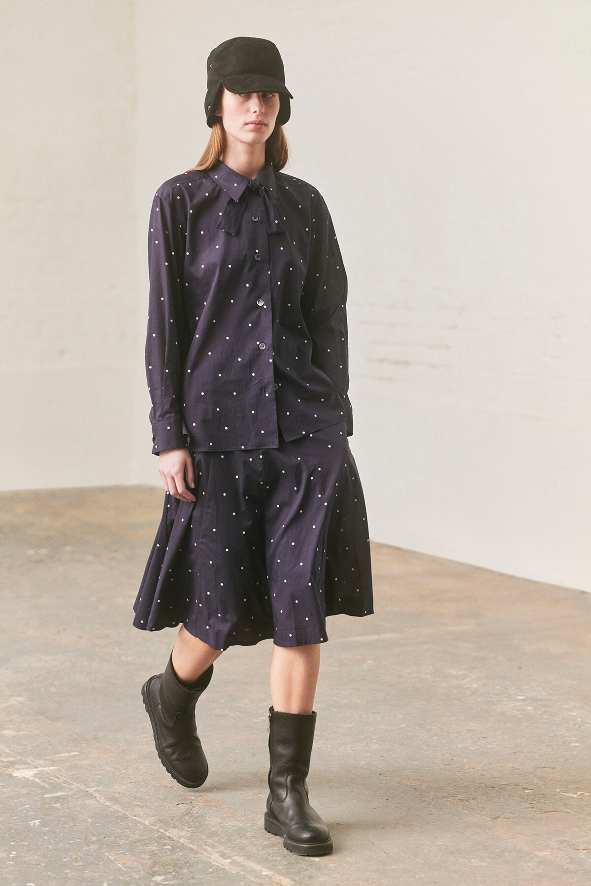 Fashion model wearing a navy polka-dot blouse and matching skirt, with a black cap and chunky boots in a neutral studio setting.
