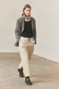 Model walking indoors in a gray blazer over a black sweater, with a beige turtleneck/scarf and beige wide-leg trousers, black boots.