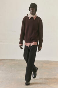 Male model wearing a burgundy knit sweater over a pink shirt, with a beige scarf, black pants and boots, walking in a minimalist studio.