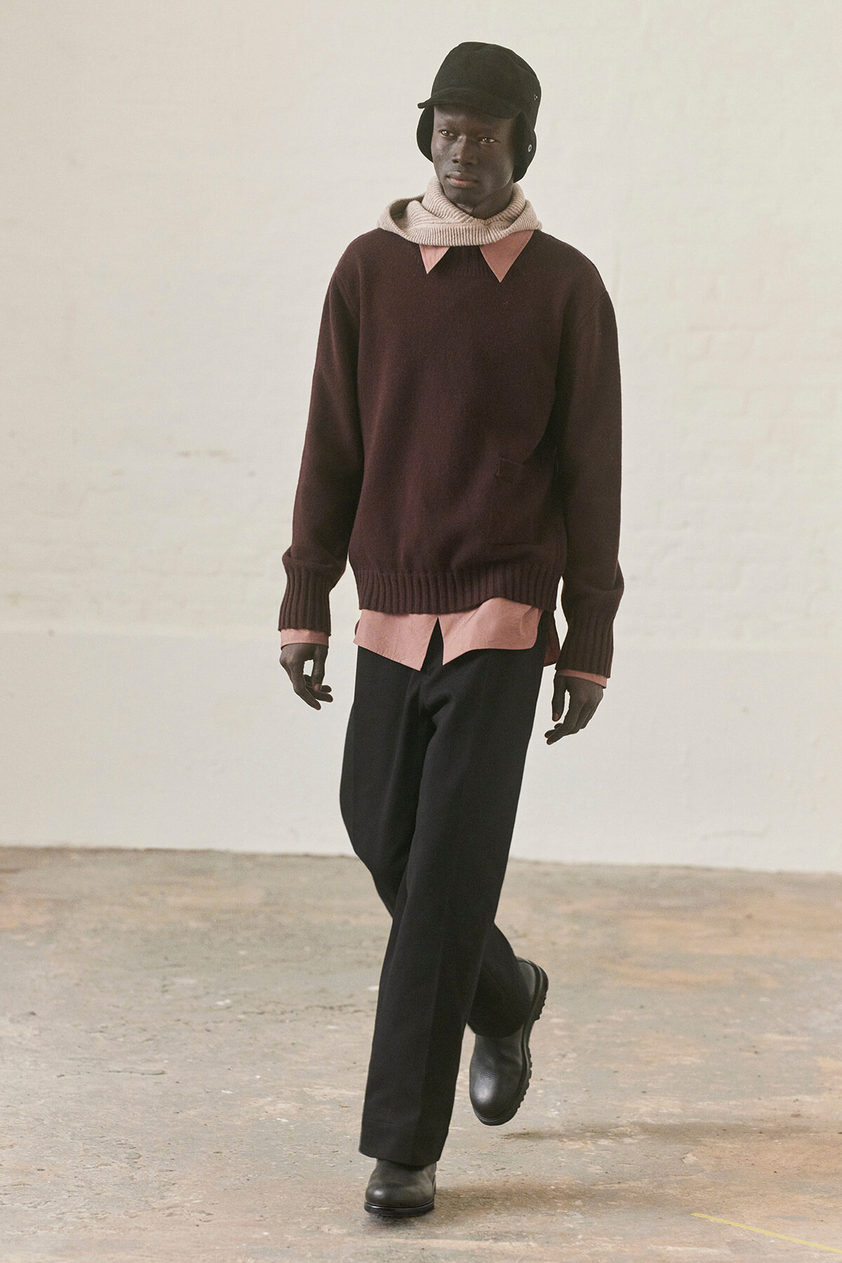 Male model wearing a burgundy knit sweater over a pink shirt, with a beige scarf, black pants and boots, walking in a minimalist studio.
