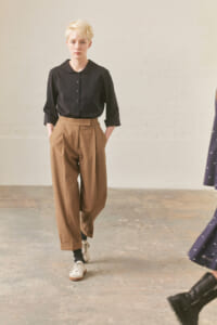 Model with short blonde hair wearing a black button-down blouse and tan high-waisted wide-leg pants with pockets, walking toward the camera in a studio.