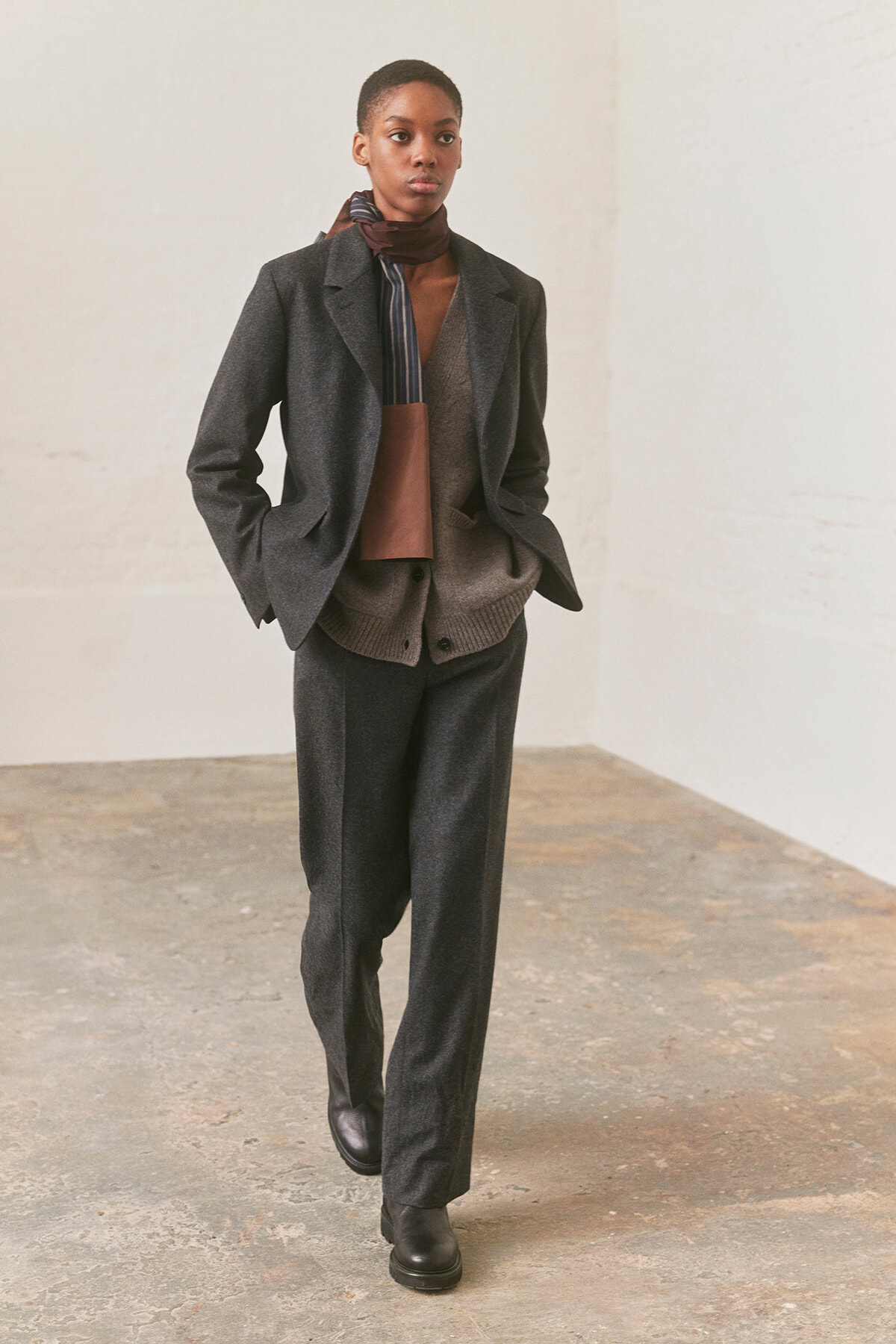 Person wearing a charcoal blazer, matching trousers, and a knit cardigan with a brown scarf in an empty studio.