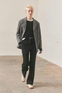 Person with short blonde hair wearing a gray blazer, black sweater, and black trousers, walking indoors toward the camera.