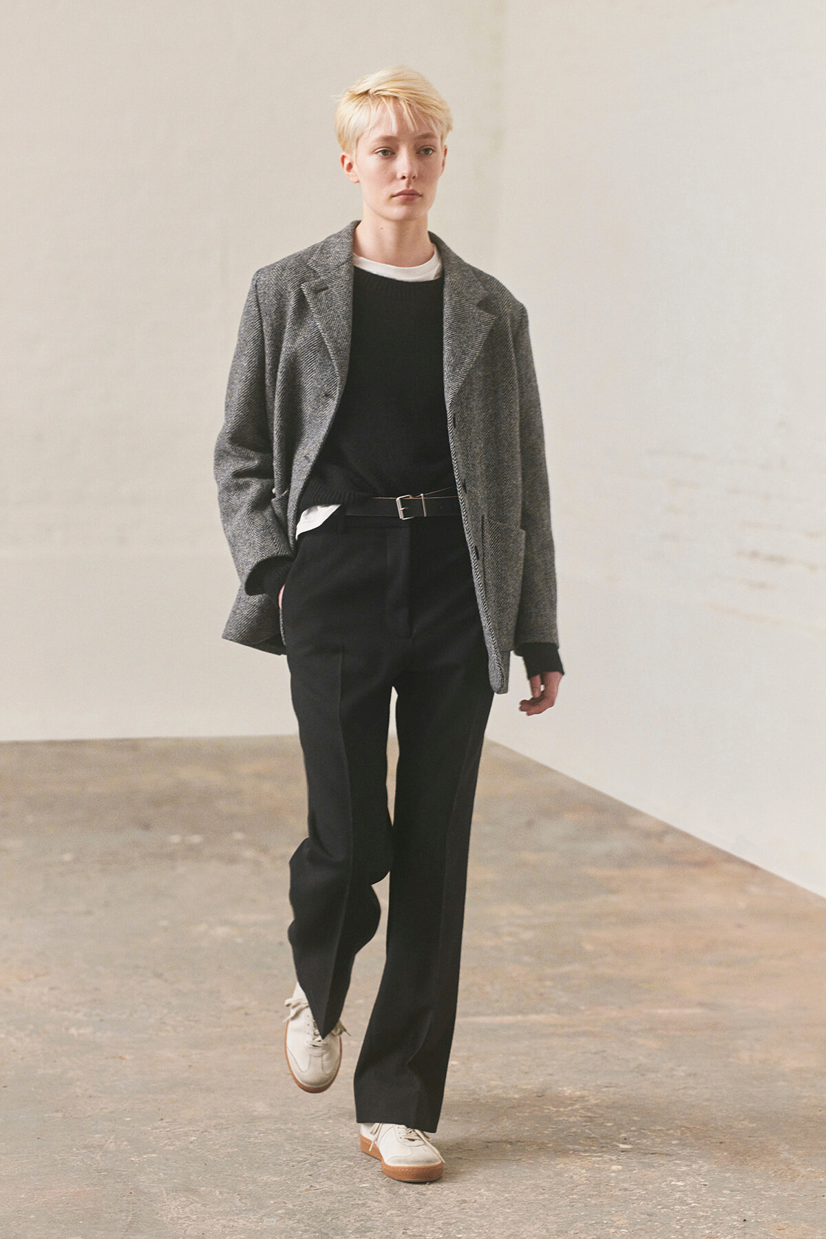 Person with short blonde hair wearing a gray blazer, black sweater, and black trousers, walking indoors toward the camera.