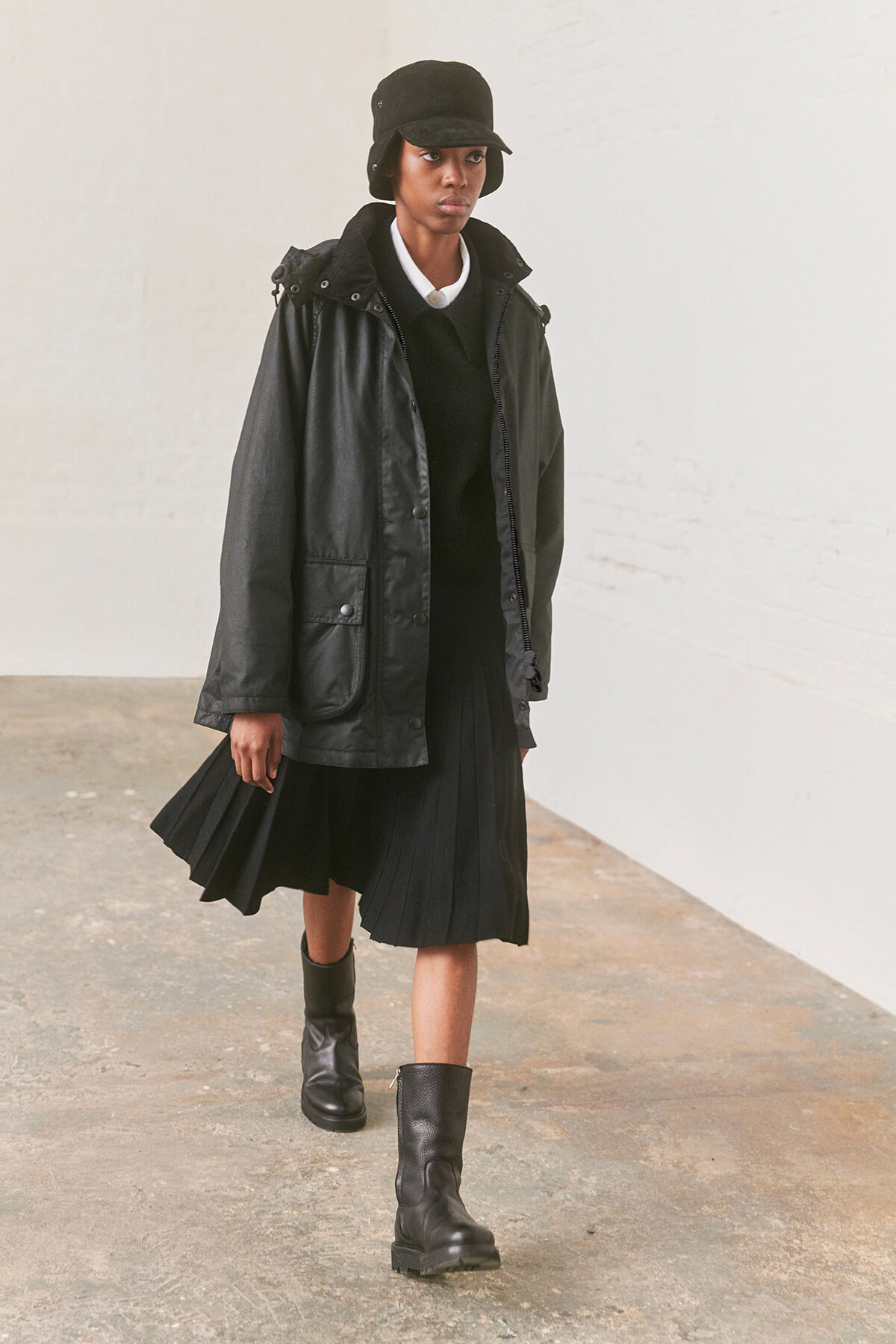 Person walking toward camera in a black hooded jacket, pleated black skirt, black boots, and black hat in an industrial setting.”,