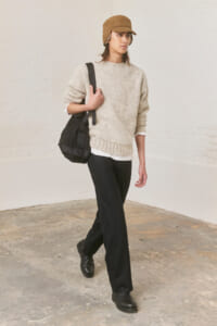 Person walking indoors in beige knit sweater, black pants, brown cap, and black backpack/duffle bag over the shoulder.