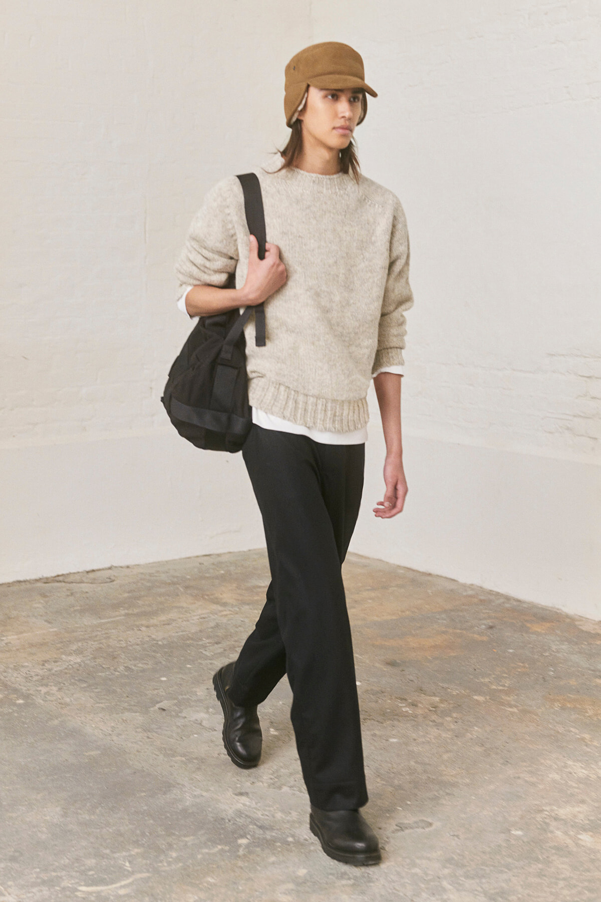 Person walking indoors in beige knit sweater, black pants, brown cap, and black backpack/duffle bag over the shoulder.