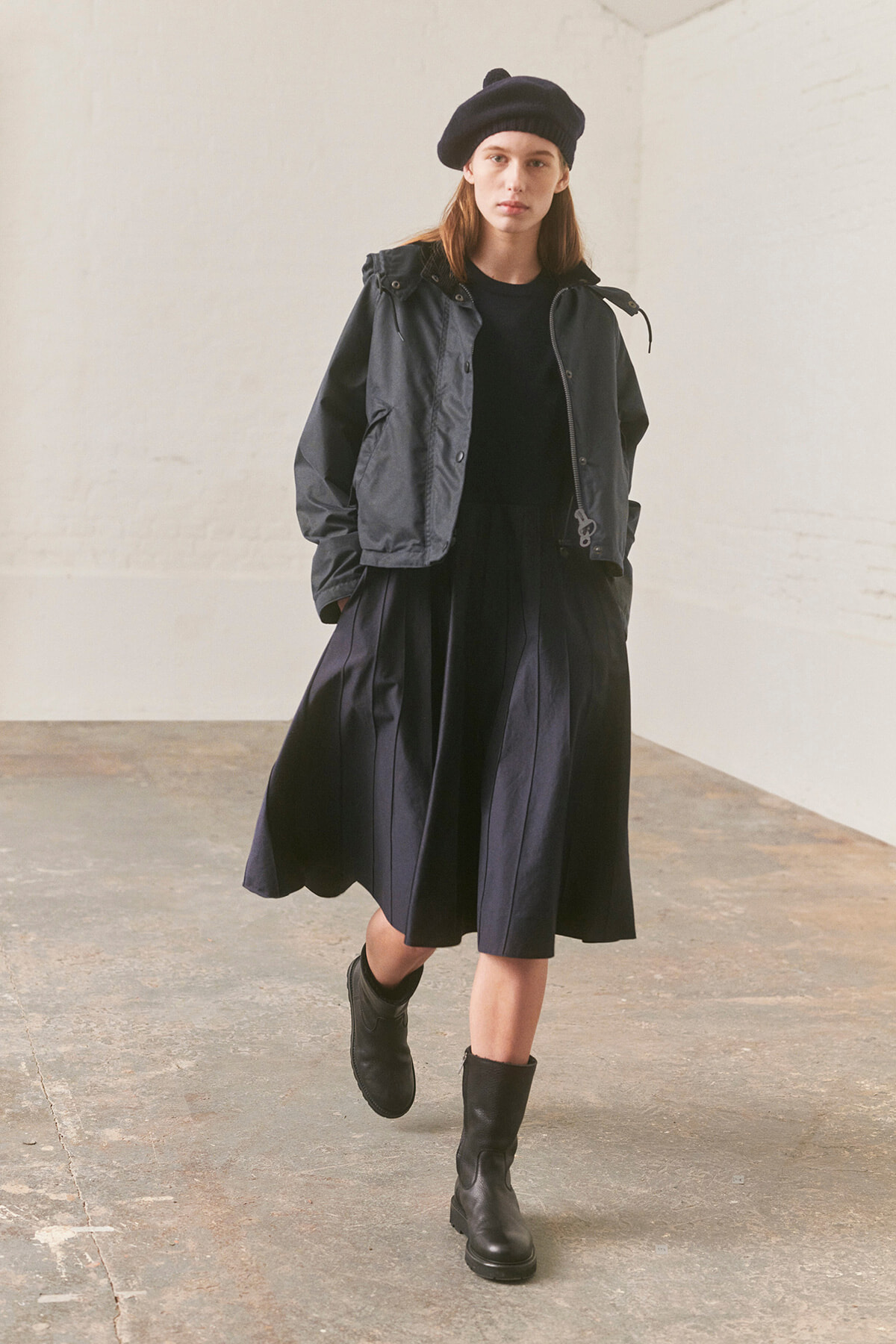 Model walking toward the camera in a black beret, oversized black jacket, and a full black skirt/dress; industrial studio setting.