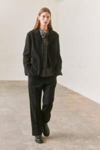 Woman in a black tailored suit with a gray ruffled blouse walking toward the camera in a minimal studio setting.