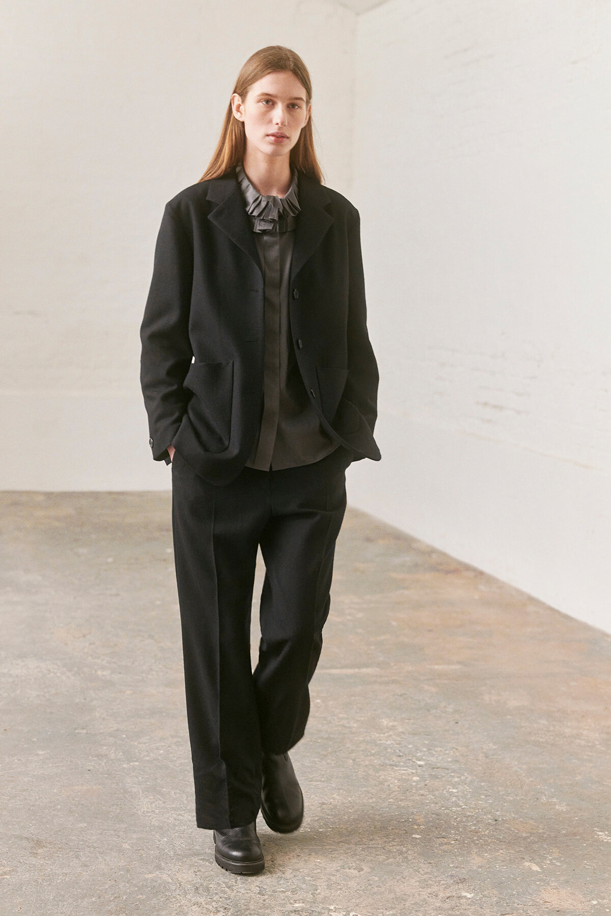 Woman in a black tailored suit with a gray ruffled blouse walking toward the camera in a minimal studio setting.