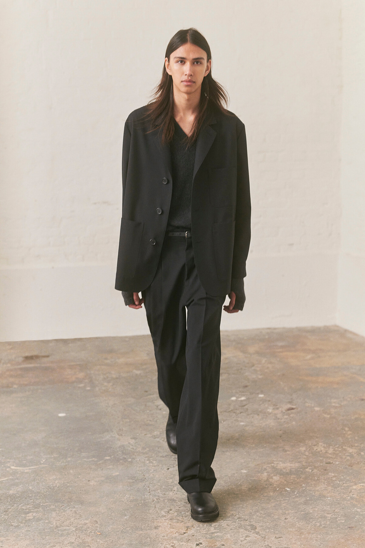 Model wearing an all-black suit (blazer and trousers) walking toward the camera in a minimalist, industrial-style room with a concrete floor.
