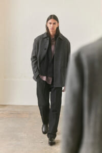 Person with shoulder-length dark hair walks toward camera in a minimalist studio, wearing a gray coat over a dark vest and mauve shirt with black trousers and black shoes.