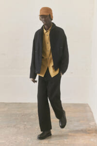 Male model wearing a mustard button-up shirt under a dark jacket, black wide-leg pants, and a brown hat with black boots in a studio setting.