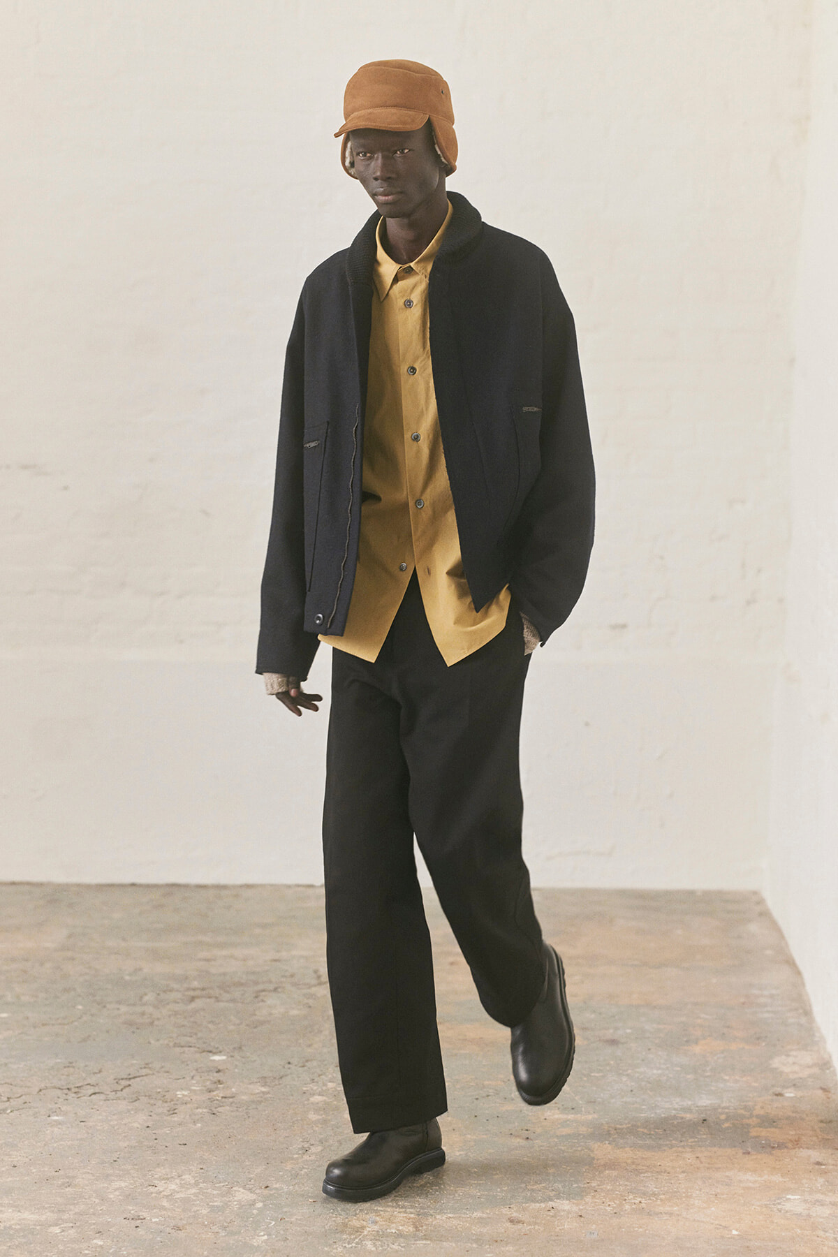 Male model wearing a mustard button-up shirt under a dark jacket, black wide-leg pants, and a brown hat with black boots in a studio setting.