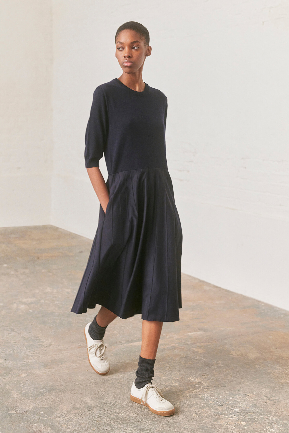 Woman in a navy three-quarter sleeve top and a full navy pleated midi skirt, standing with hands in pockets in a light studio.