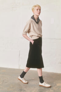 Model walking in a beige knitted sweater, black midi skirt, and white sneakers with dark socks, wearing a black patterned scarf.