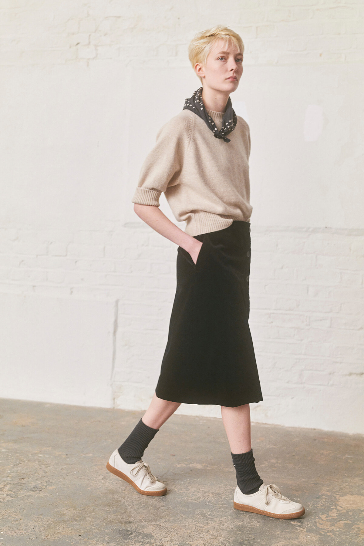 Model walking in a beige knitted sweater, black midi skirt, and white sneakers with dark socks, wearing a black patterned scarf.